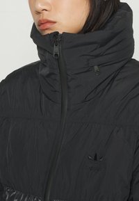 Svart vadderad jacka med hög krage, med slätt, texturerat tyg, frontdragkedja och en subtil broderad logotyp på bröstet.