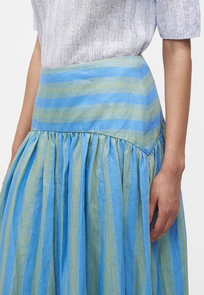 Femme portant une jupe taille haute avec des rayures horizontales et verticales bleues et vertes, associée à un top tricoté blanc à manches courtes.