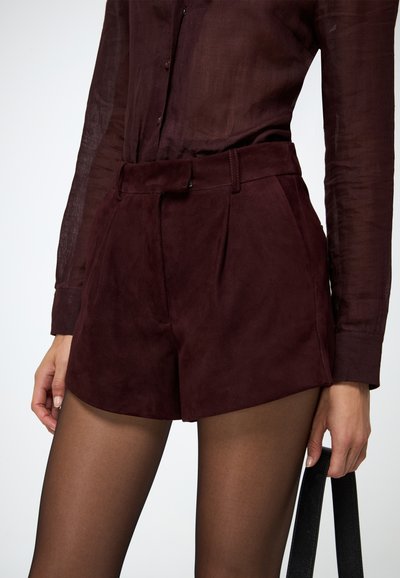 Shorts in suede color bordeaux con alta vita, frontale plissettato e passanti per cintura. Abbinati a una camicia trasparente scura con bottoni.