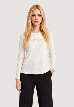 Kobieta z długimi blond włosami, ubrana w białą, długorękawową koszulę Armani Exchange i czarne spodnie, stojąca z jedną ręką w kieszeni.