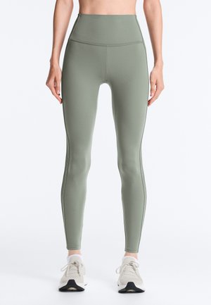 Kobieta w wysokiej talii oliwkowych legginsach i białych sneakersach, stojąca z ramionami swobodnie opuszczonymi przy bokach na jednolitym tle.