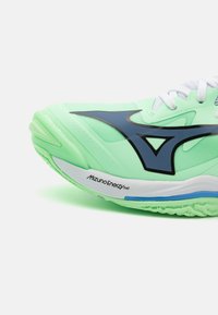 Světle zelená sportovní bota s texturovaným povrchem, modrými akcenty, bílými tkaničkami a výrazným logem Mizuno Energy na podrážce.
