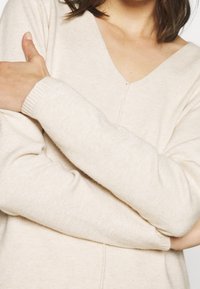 Maglione beige lavorato a maglia con scollatura a V e polsini a costine. Texture morbida con un leggero motivo mélange, mostrato con le braccia incrociate sul corpo.