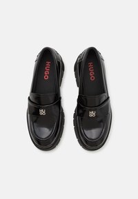 Mocassins noirs en vernis avec un bout arrondi, semelle en caoutchouc épaisse, et une sangle à l'avant avec un logo "HUGO" en argent. L'intérieur présente un logo rouge.