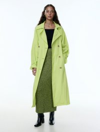Limoengroene trenchcoat met grote knopen, gecombineerd met een gestructureerde groene gebreide rok en zwarte enkelhoogte laarsjes op een witte achtergrond.