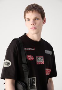 T-shirt en coton noir avec divers logos et patchs imprimés colorés, à manches courtes et col rond, porté avec un sac bandoulière.