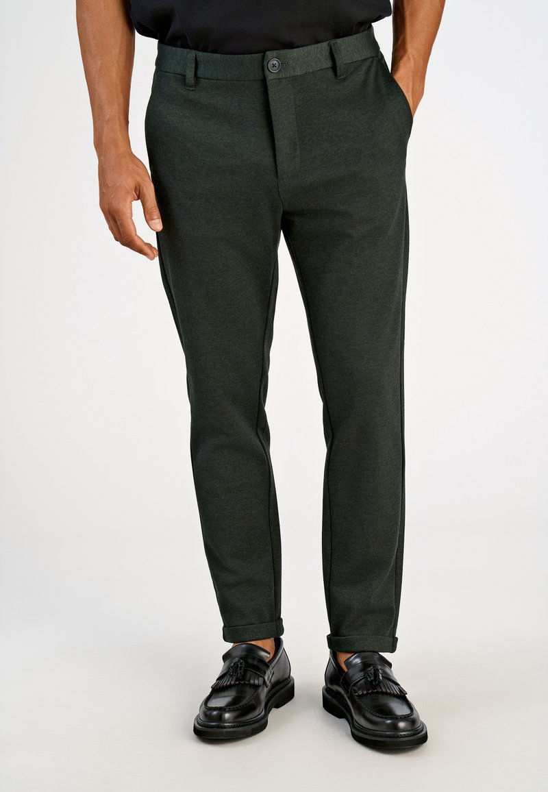 Homme portant un pantalon slim vert foncé avec des revers, associé à des mocassins en cuir noirs à pampilles et une chemise noire.