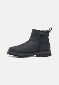 Bottine noire à la cheville avec bout rond, fermeture éclair latérale, sangle en velcro, languette au dos et semelle épaisse crantée sur fond uni.