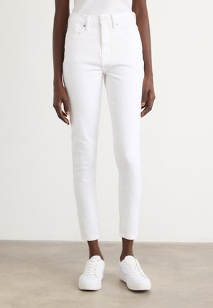 Persoon draagt witte skinny jeans met hoge taille en witte lage sneakers, staand op een lichtgrijze vloer.