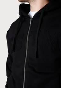 Schwarze Zip-Hoodie mit einem strukturierten, gestickten Logo. Verfügt über Kordelzüge und einen metallischen Reißverschluss. Weicher Stoff mit einer glatten Oberfläche.