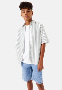 Camisa de botones a rayas en gris claro con rayas verdes, usada sobre una camiseta blanca, combinada con pantalones cortos de mezclilla azul claro. Ajuste casual.