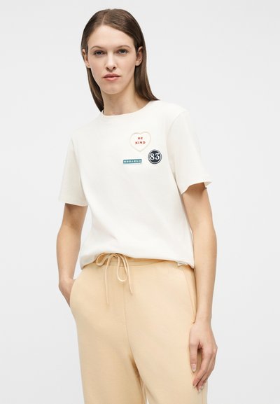 Γυναίκα που φοράει λευκό μπλουζάκι με μπαλώματα "ΝΑ ΕΙΣΑΙ ΚΑΛΟΣ", "ΣΑΒΒΑΤΟΚΥΡΙΑΚΟ" και "83", συνδυασμένο με μπεζ παντελόνια με κορδόνι, στέκεται μπροστά σε λευκό φόντο.