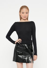 Donna con capelli lisci e chiari, che indossa un top nero a maniche lunghe e una mini gonna nera lucida con uno spacco frontale, posando su uno sfondo bianco a tinta unita.