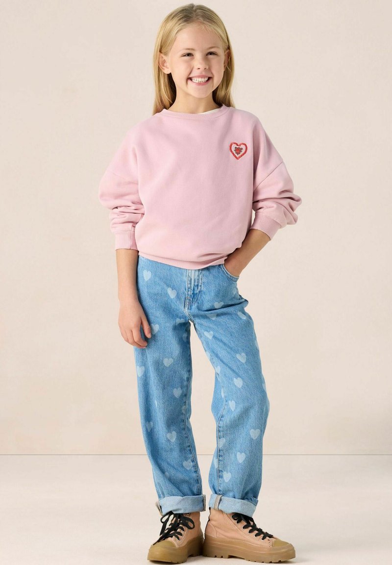 Sweatshirt rose avec un patch en forme de cœur, jean en denim bleu clair ornés de motifs en forme de cœur, et chaussures beiges à lacets avec une semelle épaisse.