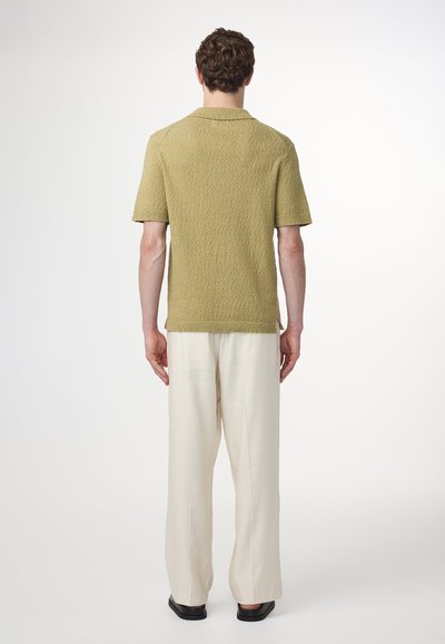 Korte mouwen polo in gestructureerd olijfgroen breisel, gecombineerd met ruime off-white broeken en zwarte schoenen, gezien vanaf de achterkant.