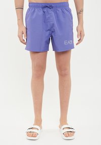 Boxer da piscina viola con elastico in vita e cordino regolabile. Presenta un logo riflettente EA7 sul lato. Abbinato a sandali bianchi.