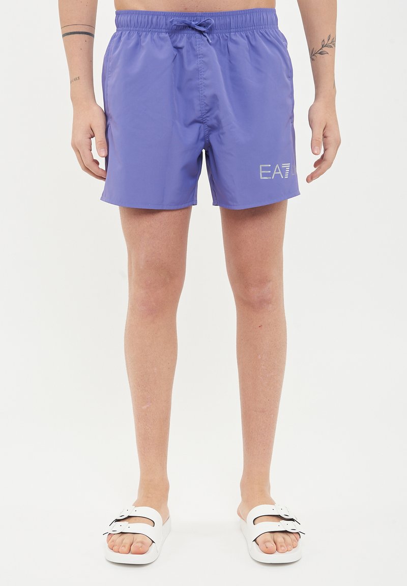 Boxer da piscina viola con elastico in vita e cordino regolabile. Presenta un logo riflettente EA7 sul lato. Abbinato a sandali bianchi.