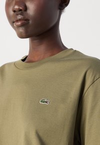 Olivenfarbene Baumwoll-T-Shirt mit Rundhalsausschnitt und kurzen Ärmeln, mit einem kleinen gestickten Krokodil-Logo auf der linken Brust.