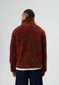 Veste en polaire rouge et marron à motifs avec une finition texturée, dotée d'un col montant et de formes en diamant. Manches mi-longues et coupe décontractée.