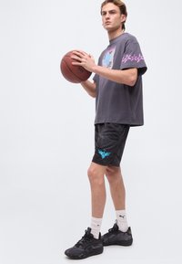 Νεαρός άνδρας κρατάει ένα μπάσκετ, φοράει γκρι μπλουζάκι με σχέδιο, μαύρο σορτς με μοτίβο και μπλε λογότυπο, λευκές κάλτσες Puma και μαύρα αθλητικά παπούτσια.