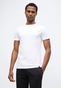 Fiatal férfi sima fehér, rövid ujjú pólóban és fekete nadrágban áll, egyik kezével a zsebében, fehér háttér előtt.