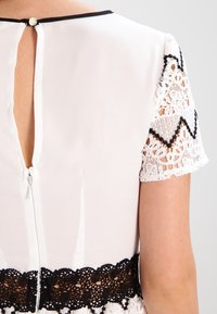 Vit klänning med svarta spetsdetaljer vid ärmarna och fållen, med en nyckelhålöppning bak och dragkedjestängning.