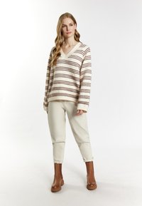 Jonge vrouw staat in een crèmekleurige gestreepte V-hals trui, lichtbeige opgerolde broek en bruine enkellaarzen tegen een witte achtergrond.