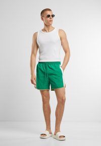 Shorts de bain verts avec des panneaux latéraux blancs et une bordure dorée, associés à un débardeur blanc et des lunettes de soleil rondes, portés avec des sandales légères.