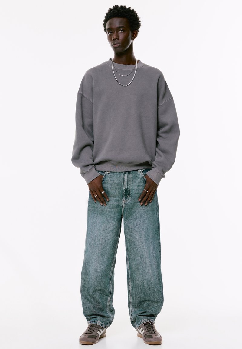 Sivi dukserica s niskim ramenima, široke svijetloplave traperice i siva tenisica. Dodaci uključuju slojevite srebrne ogrlice i prstenje.