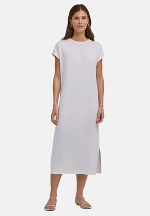 Vrouw die een effen witte midi-jurk met korte mouwen en een zijsplit draagt, met witte platte sandalen met bandjes, staand tegen een effen achtergrond.