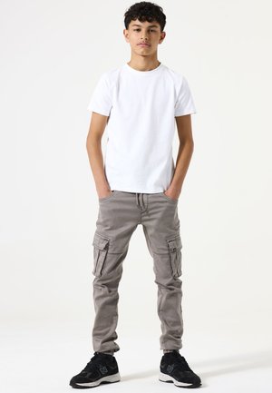 Witte T-shirt met korte mouwen gecombineerd met grijze cargobroek met zijzakken. Draagt zwarte sportschoenen, tegen een neutrale achtergrond.