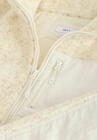 Cremefarbene, fleecegefütterte Jacke mit einem gesteppten Innenfutter, ausgestattet mit einem Doppelreißverschluss und weicher Textur. Die Außenschicht ist glatt und leicht.