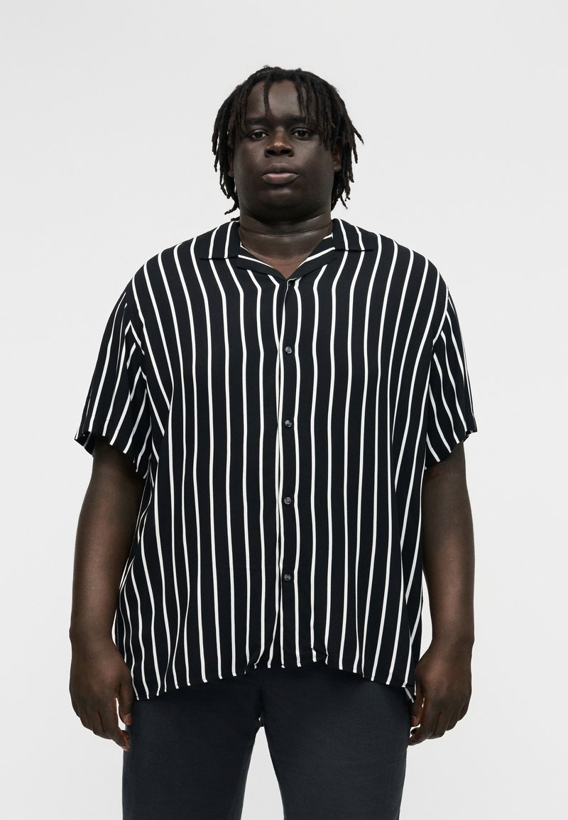 Person mit dunkler Haut und kurzen Dreadlocks trägt ein schwarz-weiß gestreiftes kurzärmliges Hemd mit Knöpfen und steht vor einem einfarbigen Hintergrund.