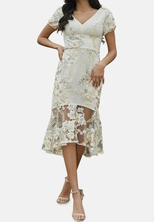 Kwiatowa sukienka haftowana w jasnym beżu z bufkami, dekoltem w kształcie litery V i przezroczystym, warstwowym dołem z detalicznymi wzorami. Sandały na ramiączkach.
