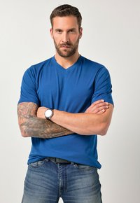 Niebieski t-shirt z krótkim rękawem i dekoltem w serek, wykonany z gładkiej tkaniny, zestawiony z dżinsami. Na ramionach widoczne są tatuaże, a na nadgarstku zegarek.