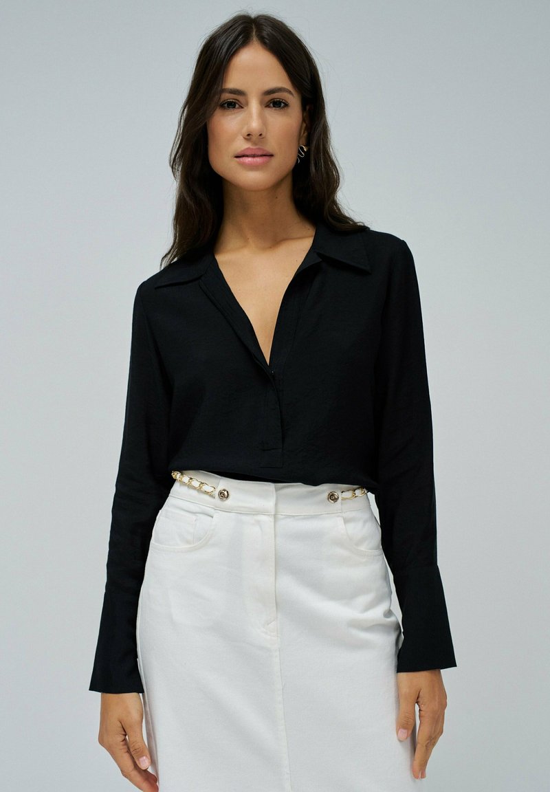 Camisa negra de manga larga con cuello, combinada con una falda blanca que presenta un detalle de cadena en la cinturilla y botones expuestos.