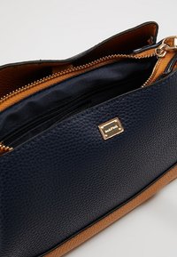 La bague en cuir texturé bleu marine avec une base beige, dotée d'une fermeture éclair et de quincaillerie dorée. L'intérieur comprend un compartiment zippé supplémentaire.