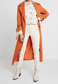 Gabardina naranja sobre una chaqueta corta color crema, combinada con pantalones color crema remangados y botas de tobillo blancas con estampado de serpiente.