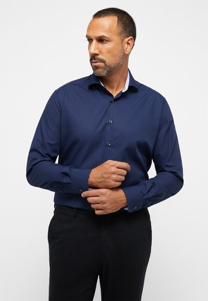 Dunkelblaues Langarmhemd mit Button-Down-Kragen, das durch kontrastierende gemusterte Manschetten und eine glatte Textur besticht; kombiniert mit dunklen Hosen.