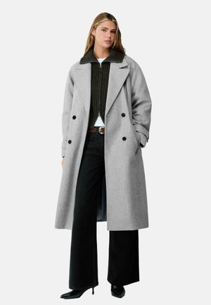 Žena má na sebe dlhý sivý dvojradový kabát cez tmavý sveter na zips, čierne nohavice s širokými nohavicami a čierne špicaté lodičky.