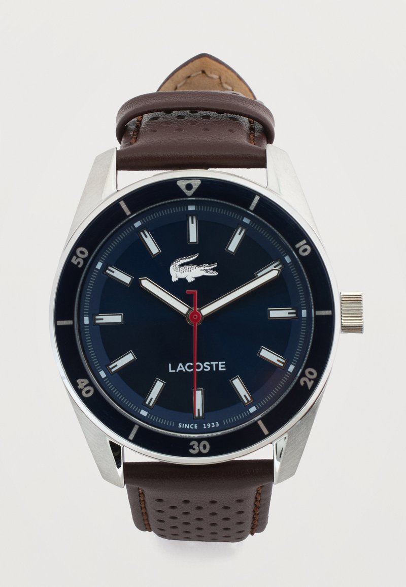 Στρογγυλό ασημένιο ρολόι με σκούρο μπλε καντράν, λευκούς δείκτες ώρας, κόκκινο δείκτη δευτερολέπτων, λογότυπο Lacoste και καφέ δερμάτινο λουράκι με τρύπες.