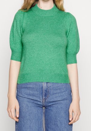 Žena v zeleném pleteném svetru s krátkými nafouklými rukávy a modrých džínách s vysokým pasem před jednobarevným pozadím.