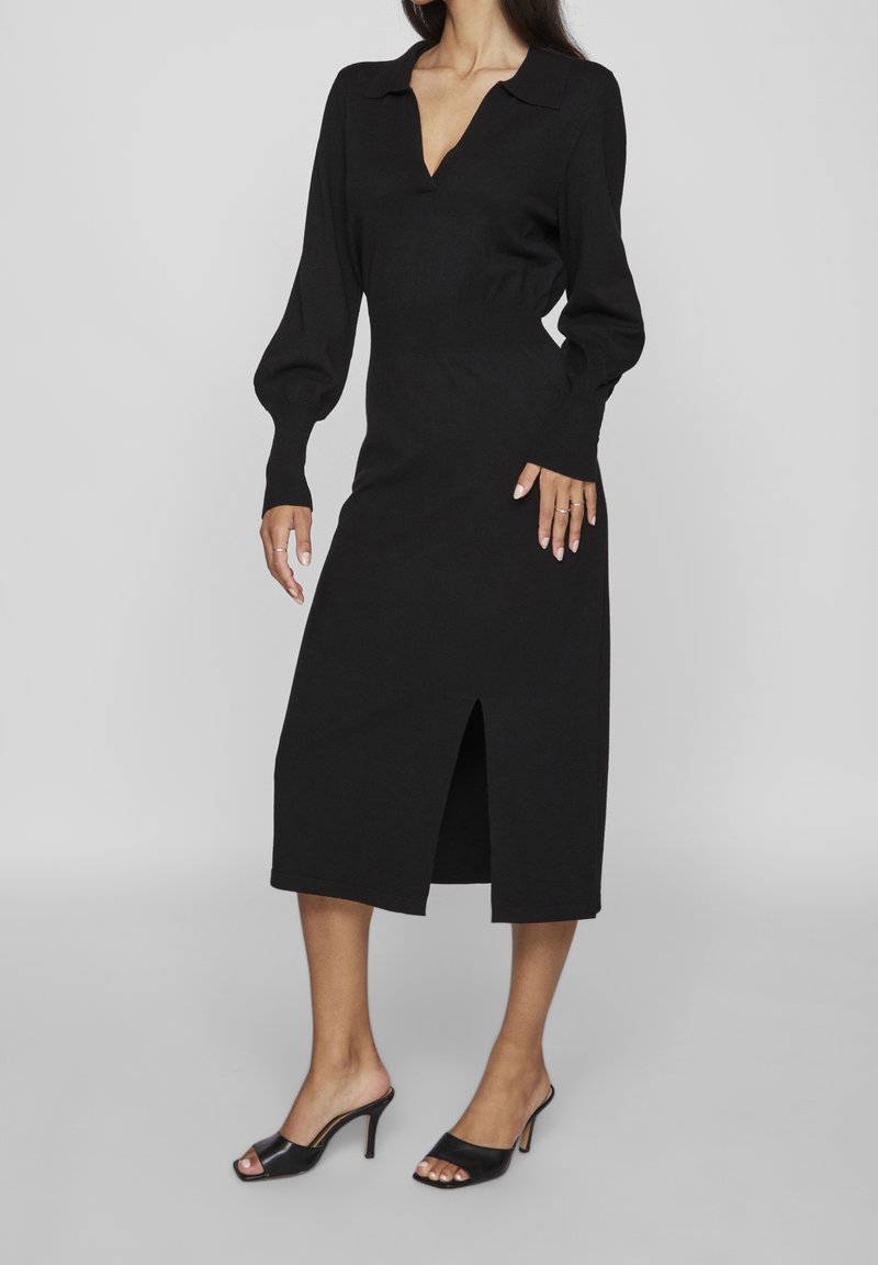 Femme portant une robe midi noire à manches longues avec un décolleté en V plongeant, un col, une fente à l'avant, et des mules à talons ouvertes noires.