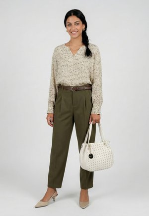 Femme aux cheveux tressés portant un chemisier léger à motifs, un pantalon taille haute olive, des talons beiges, tenant un sac à main blanc texturé, souriant sur un fond uni.