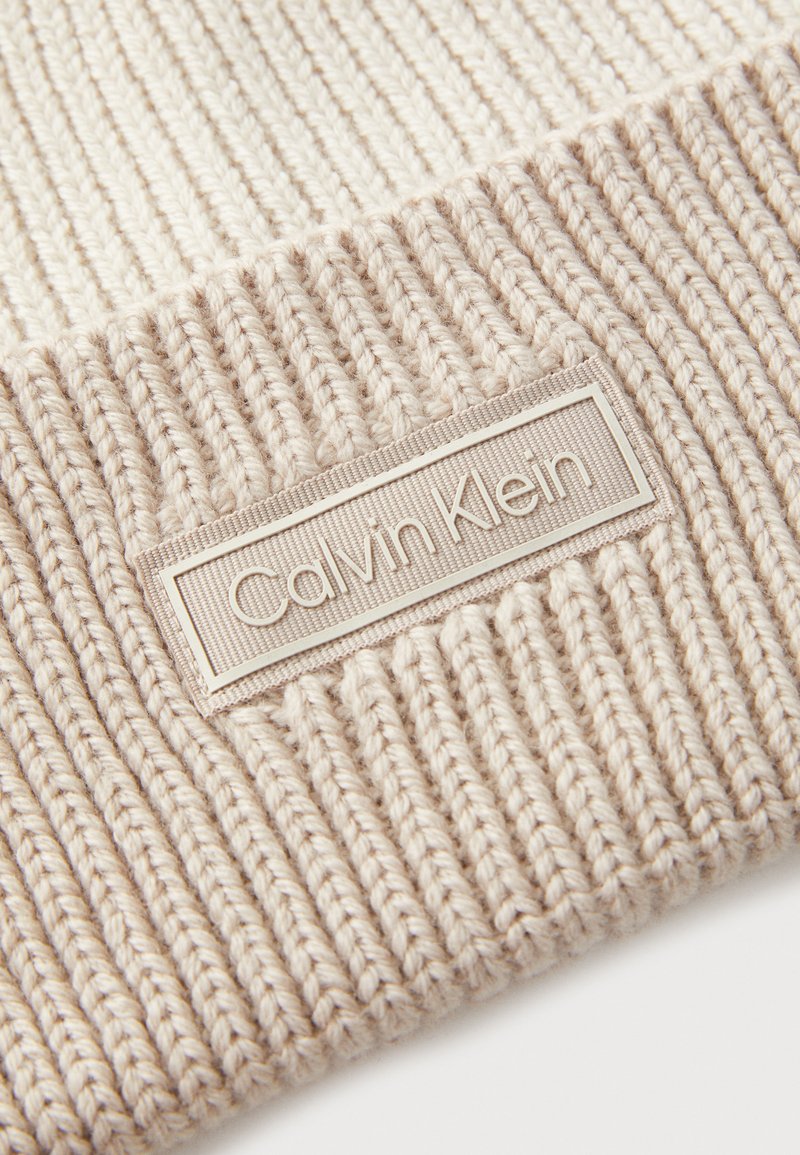 Bonnet en tricot beige avec une texture côtelée et un patch logo Calvin Klein cousu, présentant une composition en fil doux et un design sans couture.