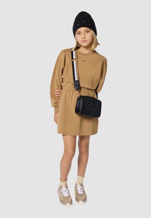 Beige langlopende jurk met elastische taille, zwarte schoudertas, zwarte gebreide muts, tan sneakers met witte veters en accenten, en gouden zolen.