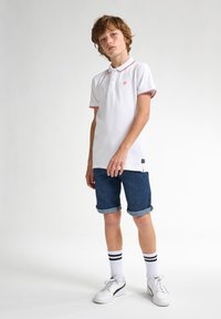 Valkoinen polo paita punaisilla yksityiskohdilla, farkkuhousut ja valkoiset lenkkarit. Malli seisoo neutraalissa taustassa, esitellen asusteiden yksityiskohtia selkeästi.