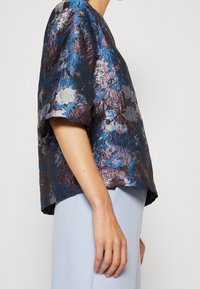 Kurze Ärmel Bluse aus strukturiertem, metallischem Stoff mit blauen und lila Blumenmustern, kombiniert mit einem hellblauen, fließenden Saum.