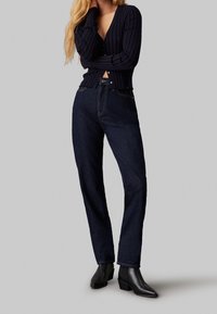 Cárdigan de punto con cuello en V, cierre de botones y dobladillo deshilachado en color marino, combinado con jeans de pierna recta de mezclilla oscura y botas negras al tobillo.