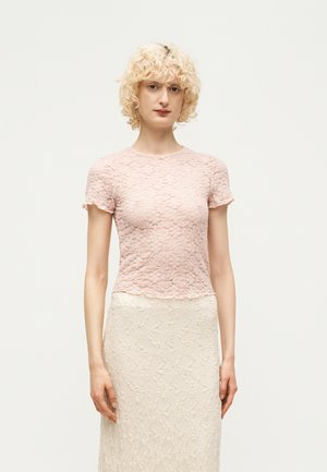 Personne aux cheveux blonds bouclés portant un haut en dentelle rose à manches courtes et une jupe en dentelle crème, debout devant un fond uni.